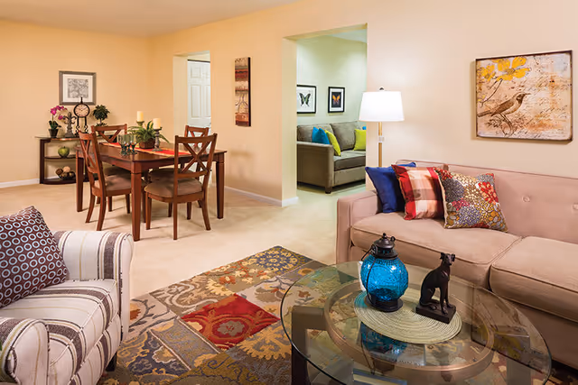 A cozy living and dining area in a senior living community. The living room features a beige sofa with colorful pillows, a striped armchair, a glass coffee table with decorative items, and a floor lamp. The dining area has a wooden table with four chairs and a side table with plants and decor. The walls are painted light beige with framed artwork, and a patterned rug covers part of the carpeted floor.