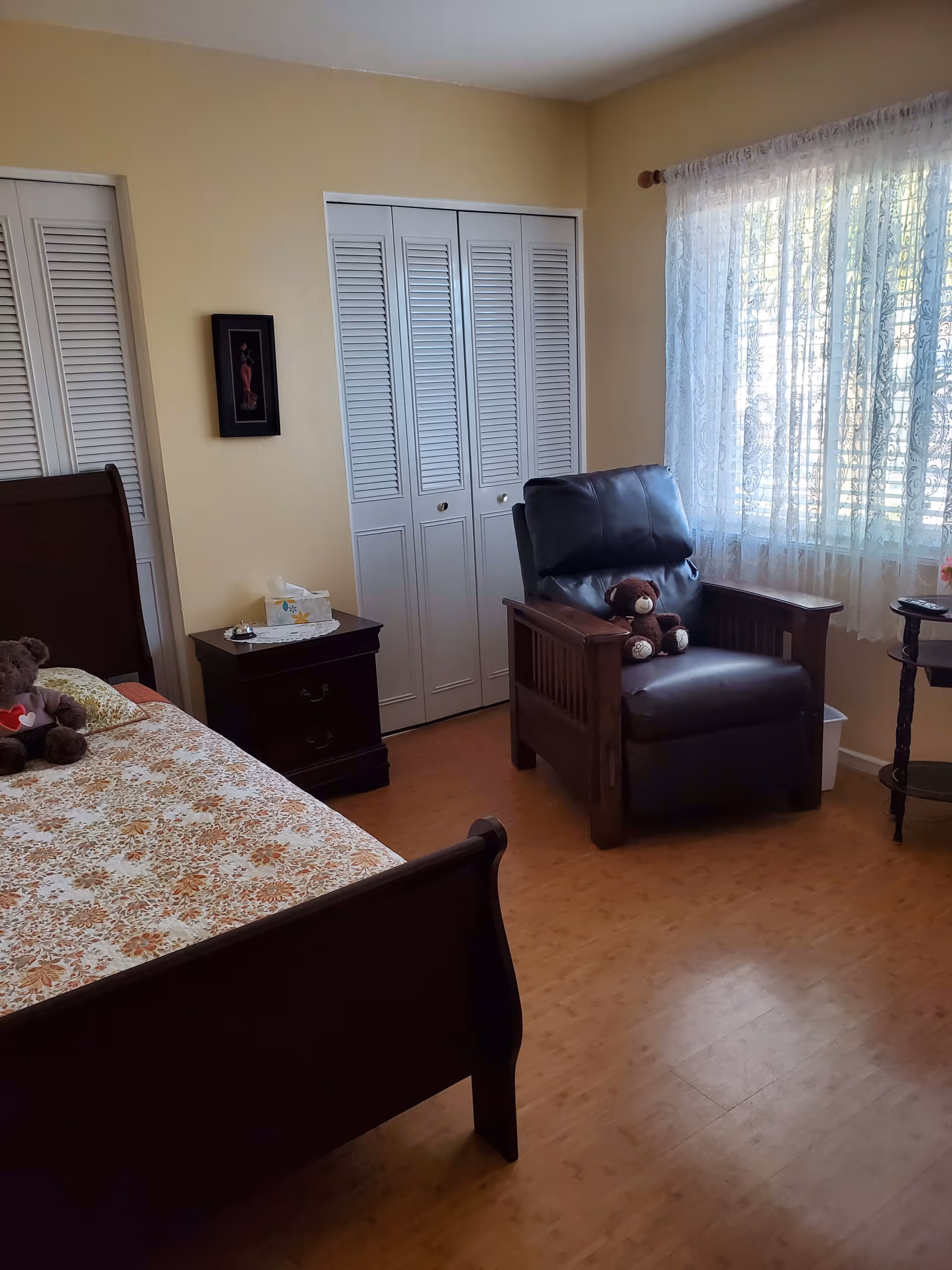 Sunlit bedroom with a bed, nightstand, closet doors, and a leather armchair by a window with lace curtains.