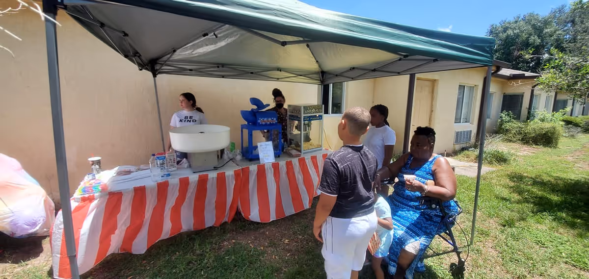 A small outdoor concession stand under a canopy tent with a red and white striped tablecloth. Two women are behind the table serving snacks including cotton candy and popcorn. In front of the stand, a woman in a blue dress sits on a walker seat while two children stand nearby. The setting is outside a beige building with grass and trees around.