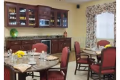 Dining area with round tables set for meals, red cushioned chairs, and a kitchenette with dark wood cabinets, a microwave, and a granite countertop. The room has light-colored walls and a window with floral curtains.