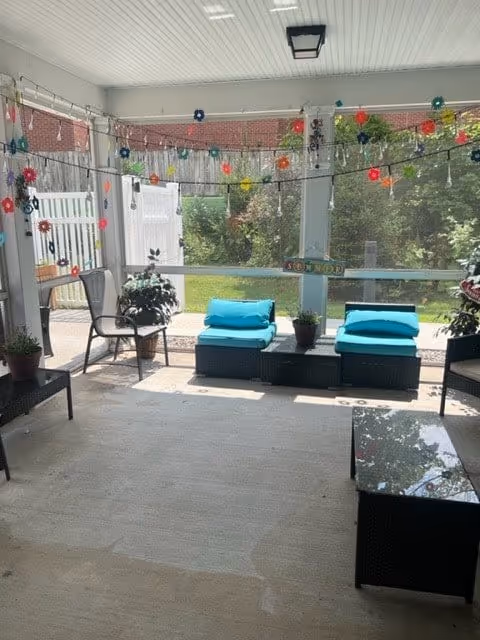 A covered outdoor patio area with wicker furniture including chairs and cushioned seats with turquoise pillows. The patio is decorated with colorful hanging flower decorations and string lights. There are potted plants on the floor and a green lawn visible through the screened enclosure.