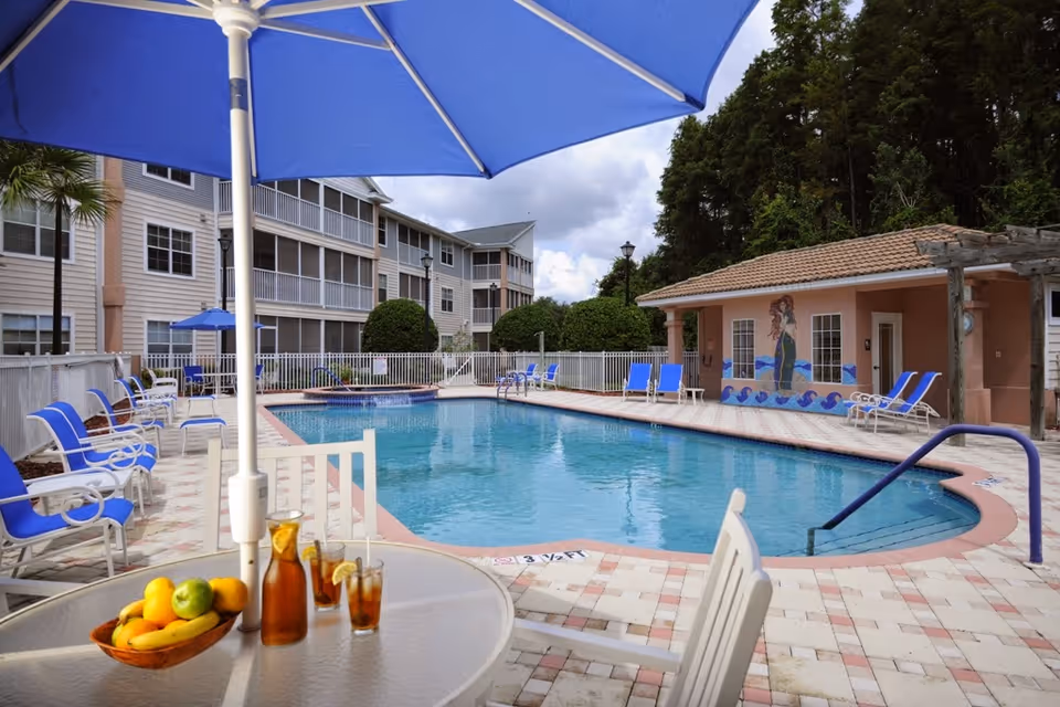 Outdoor swimming pool area at a senior living facility with blue lounge chairs, a table with a blue umbrella, a bowl of fruit, and glasses of iced tea. There is a building with a mural of a mermaid on the wall and trees in the background.