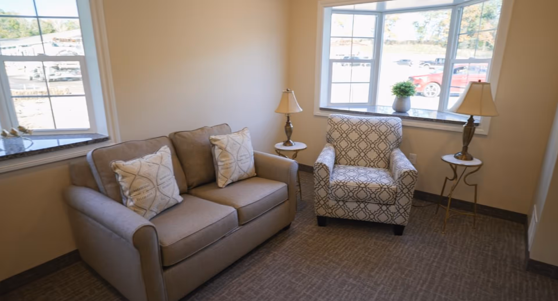 A cozy sitting area with a beige loveseat adorned with two patterned pillows, a patterned armchair, two small round side tables each holding a lamp, and two large windows letting in natural light.