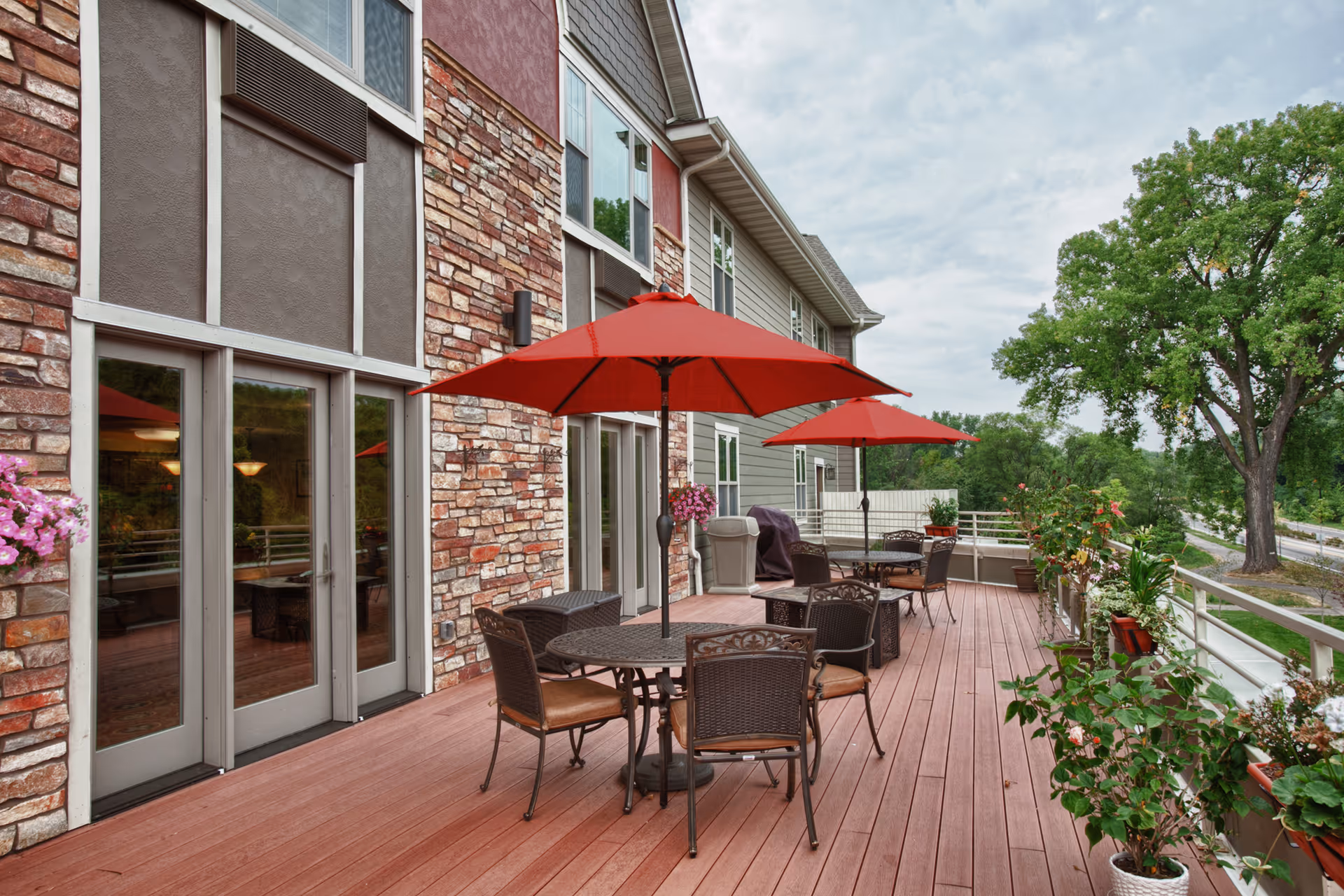 A spacious outdoor deck with red umbrellas, metal tables and chairs, potted plants, and a stone-faced building overlooking trees.