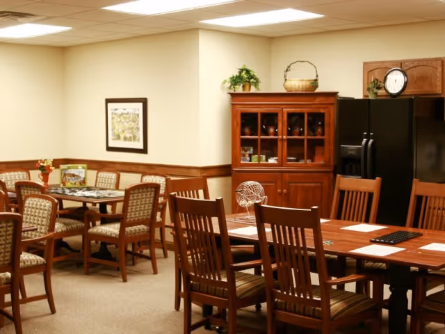 A cozy dining area with wooden tables and chairs, a wooden cabinet with glass doors displaying dishes, a black refrigerator, and a wall clock. The room has beige walls with wood paneling and a framed picture hanging on the wall.