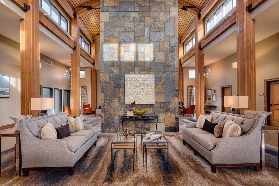 A spacious and well-lit living room area with two gray sofas facing each other, a stone accent wall in the center, and large wooden beams supporting the high ceiling. The room features modern decor including a glass coffee table, lamps on side tables, and large windows near the ceiling letting in natural light.