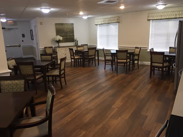 Spacious dining room with several wooden tables and upholstered chairs on a wood floor and windows with blinds.