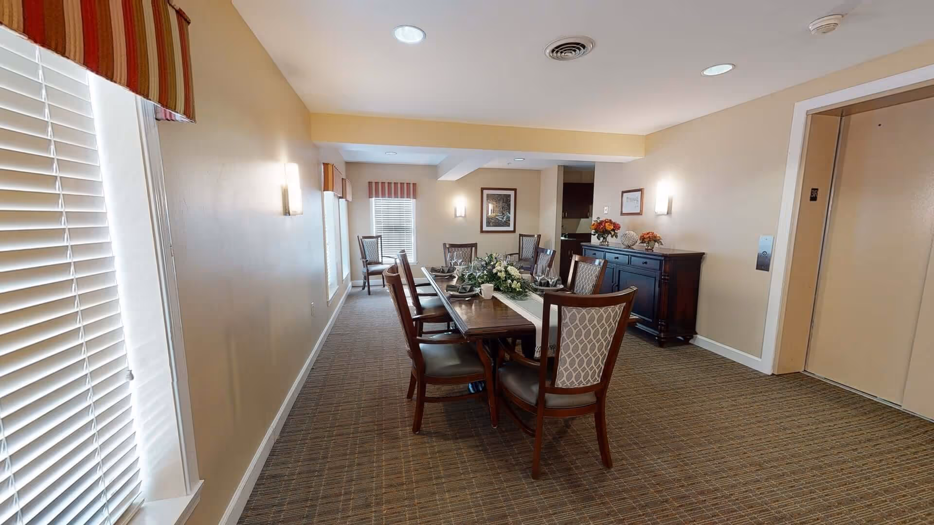 A dining room with a long wooden table set for six people, decorated with a floral centerpiece and tableware. The room has beige walls, carpeted floor, and windows with blinds and striped valances. There is a dark wooden sideboard with flower arrangements and framed pictures on the walls. An elevator door is visible on the right side of the room.