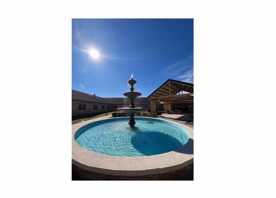 Outdoor courtyard area with a multi-tiered water fountain in the center, surrounded by a circular pool of clear water. In the background, there is a building with a covered patio area under a wooden roof structure. The sky is clear and sunny.