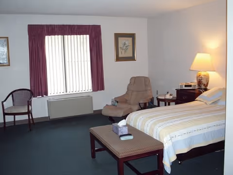 A simple bedroom with a bed covered in a striped bedspread, a cushioned armchair with an ottoman, a wooden chair, a window with vertical blinds and purple curtains, a nightstand with a lamp, and framed artwork on the walls.