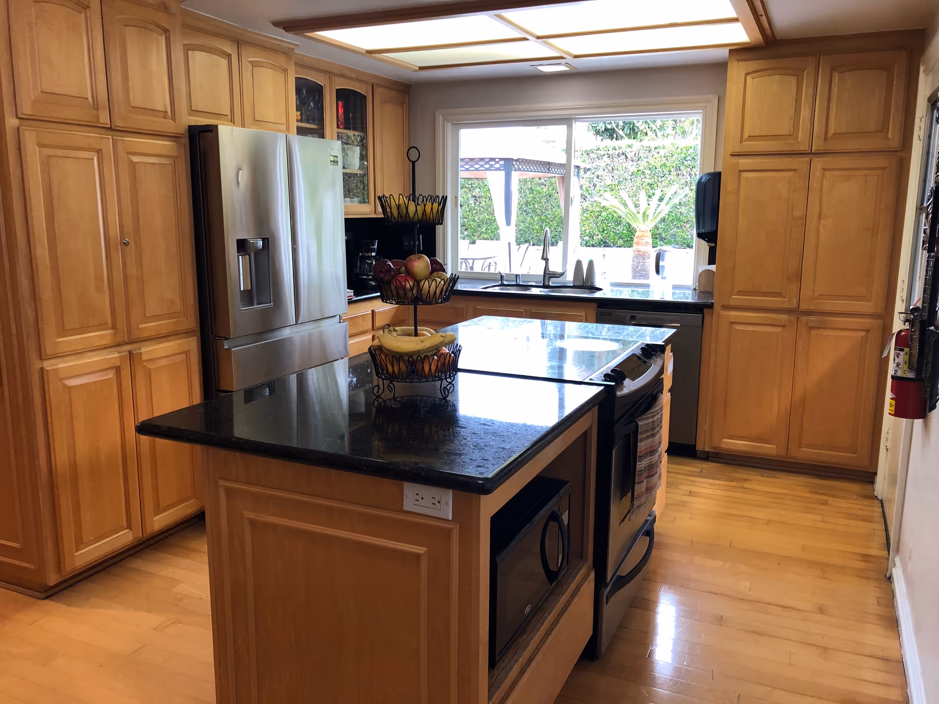 A bright kitchen with wooden cabinets and a black granite countertop island. The kitchen features a stainless steel refrigerator, a built-in microwave, an oven with a stovetop, and a dishwasher. A large window above the sink looks out onto a garden with greenery and a pergola. There is a three-tier fruit basket on the island holding bananas and apples.