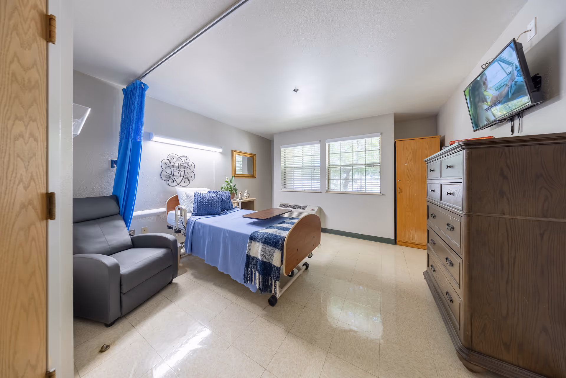 A well-lit senior living facility bedroom with a hospital-style bed covered in blue bedding and a checkered blanket. Next to the bed is a gray recliner chair and a small bedside table with a plant and decorative items. The room has two windows with blinds, a wooden dresser, a wooden wardrobe, and a wall-mounted TV showing a program. A blue privacy curtain is partially visible near the bed.