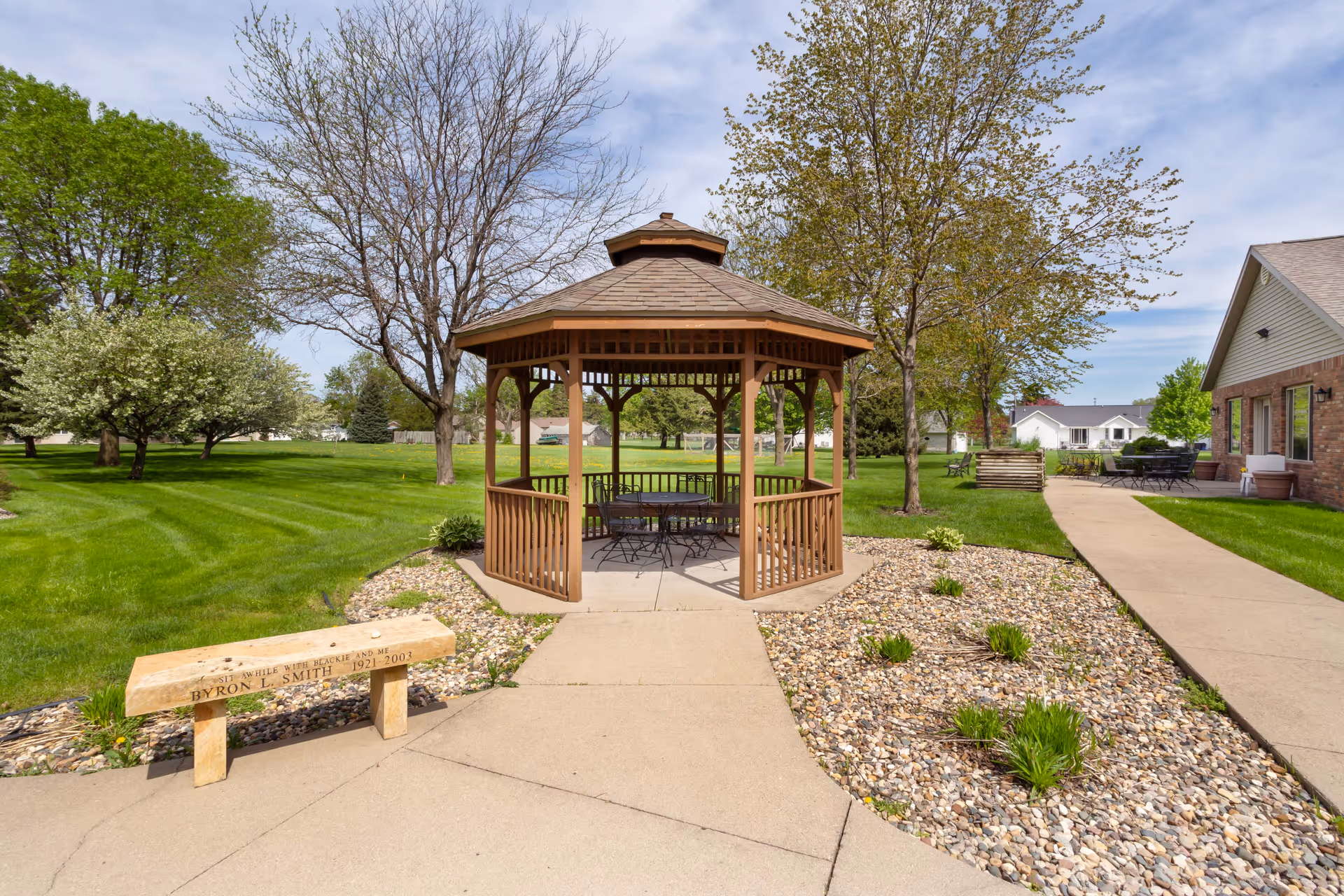 Outdoor area at Mankato Lodge Senior Living featuring a wooden gazebo with a table and chairs inside, surrounded by green grass, trees, and a stone bench with an inscription. A paved walkway leads to the gazebo and extends alongside a brick building with outdoor seating.