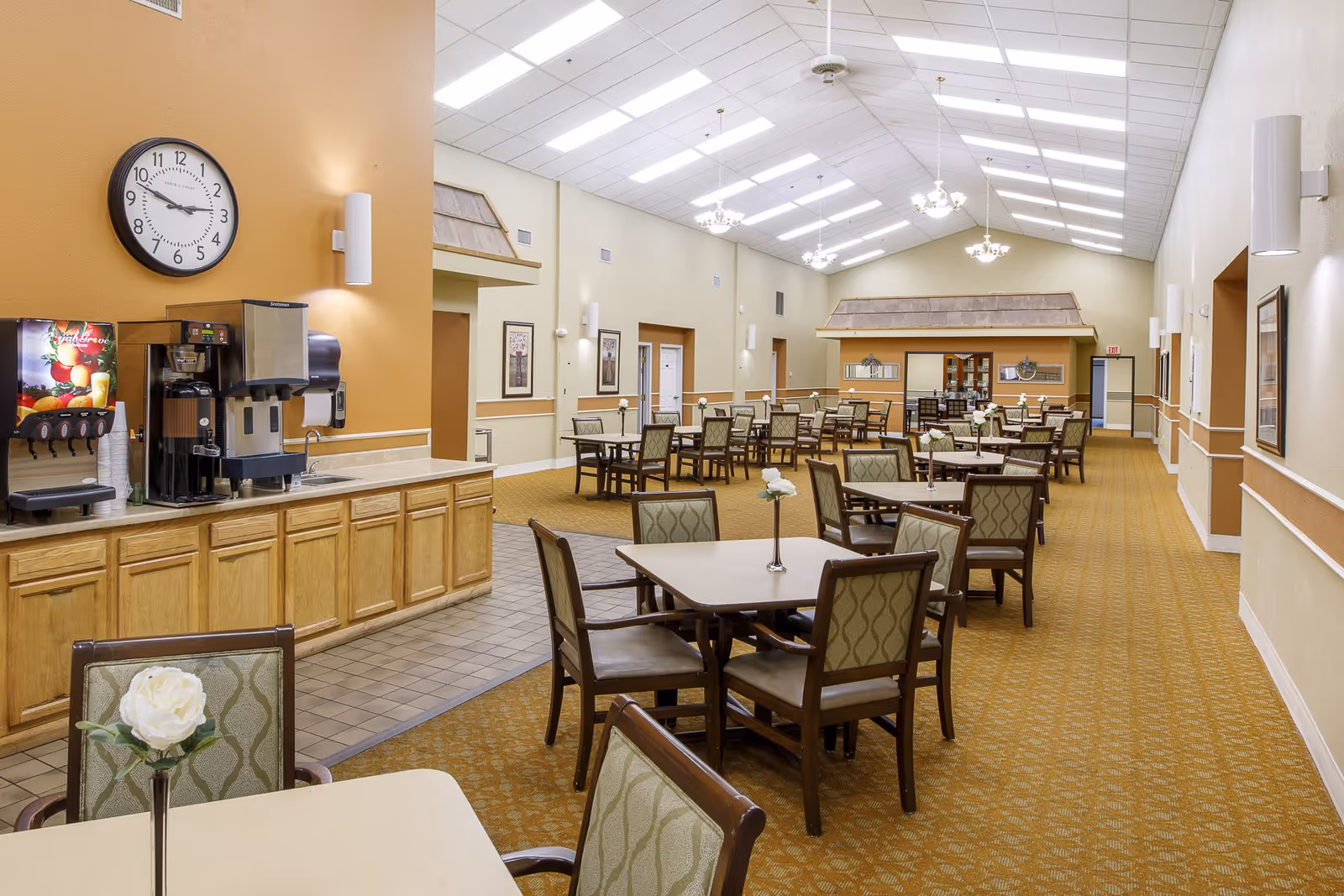 Spacious senior dining room with multiple tables and chairs, a beverage station and a vaulted ceiling with chandeliers.