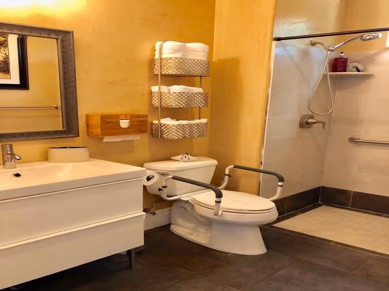 A bathroom with a white sink and vanity on the left, a toilet with safety rails in the center, and a walk-in shower with a handheld showerhead and grab bars on the right. Above the toilet, there are three metal shelves holding rolled white towels. The walls are painted a warm yellow color.