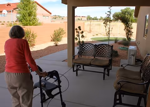 A person using a walker stands on a covered patio with outdoor seating overlooking a fenced yard.