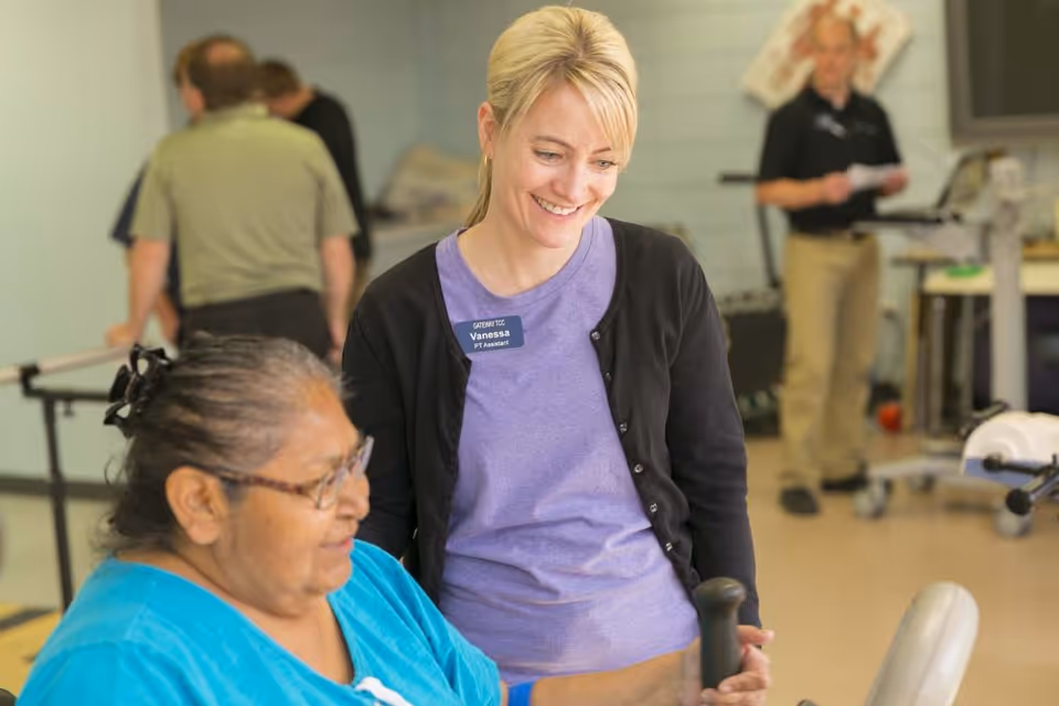 A physical therapy assistant named Vanessa is smiling and assisting an elderly woman who is using exercise equipment in a rehabilitation or therapy room. In the background, two men are engaged in activities related to physical therapy or rehabilitation.