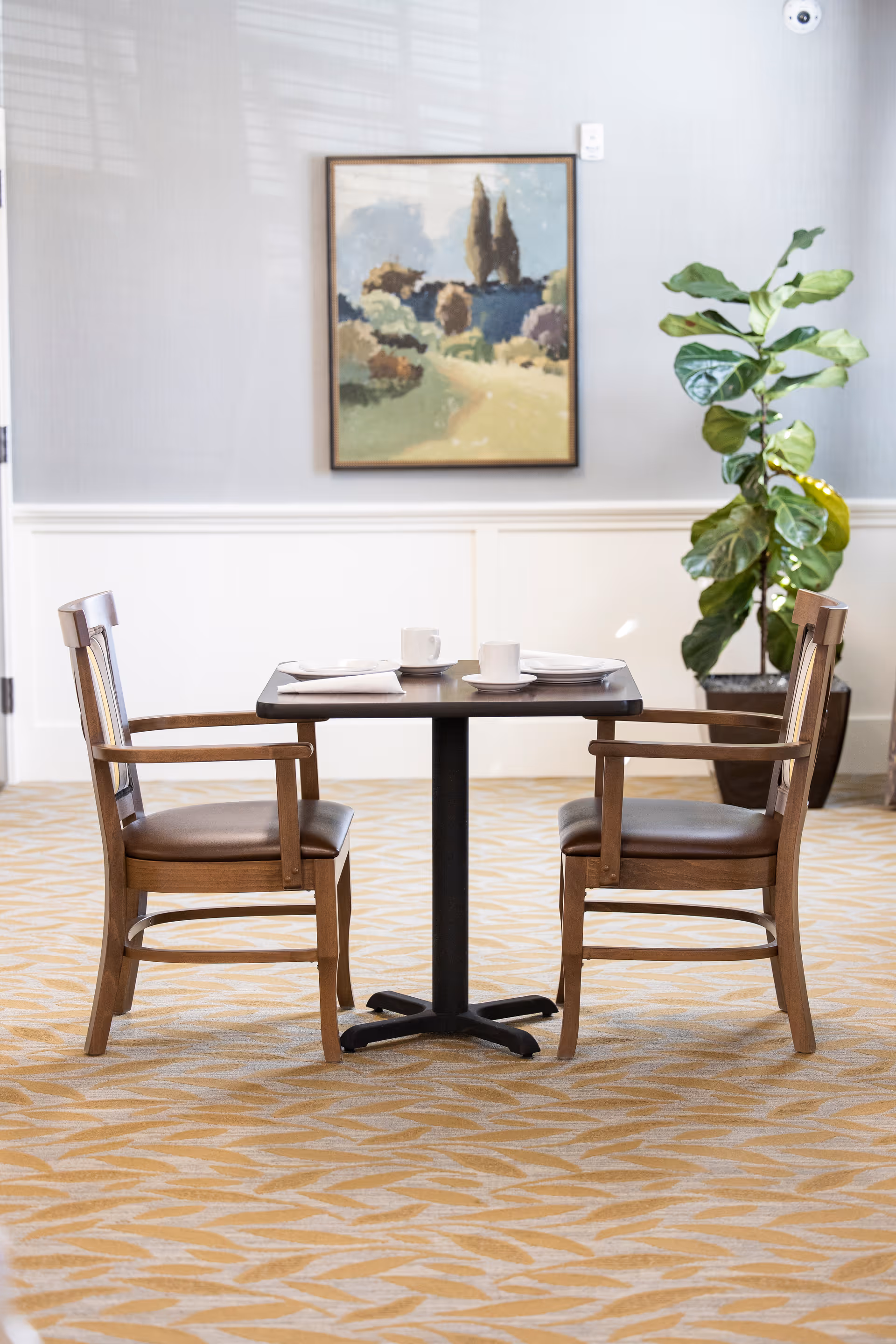 A small dining table set for two with wooden chairs in a bright dining area, with a framed landscape painting and a potted plant.