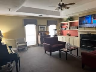 A cozy living room with beige walls and carpeted floor, featuring a ceiling fan with lights, a flat-screen TV mounted above a fireplace, built-in shelves with decorative items, a dark red armchair and loveseat, a coffee table, and a window with a valance letting in natural light.
