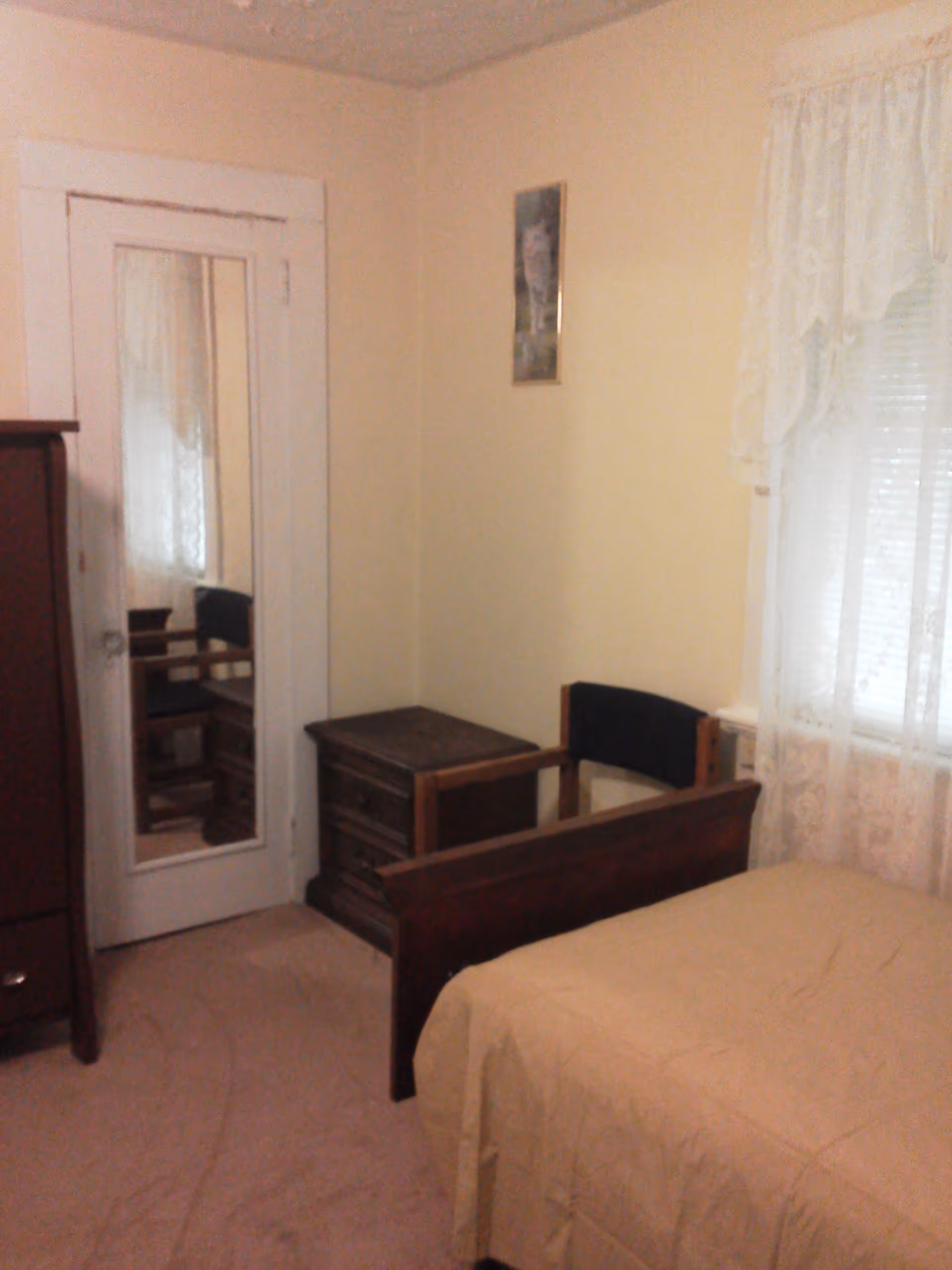 A small bedroom with beige walls and carpet. There is a wooden bed with a beige bedspread, a wooden nightstand with drawers, a wooden chair with a black cushion, and a wardrobe with a mirrored door. A window with white lace curtains is on the right wall, and a small framed picture hangs above the chair.