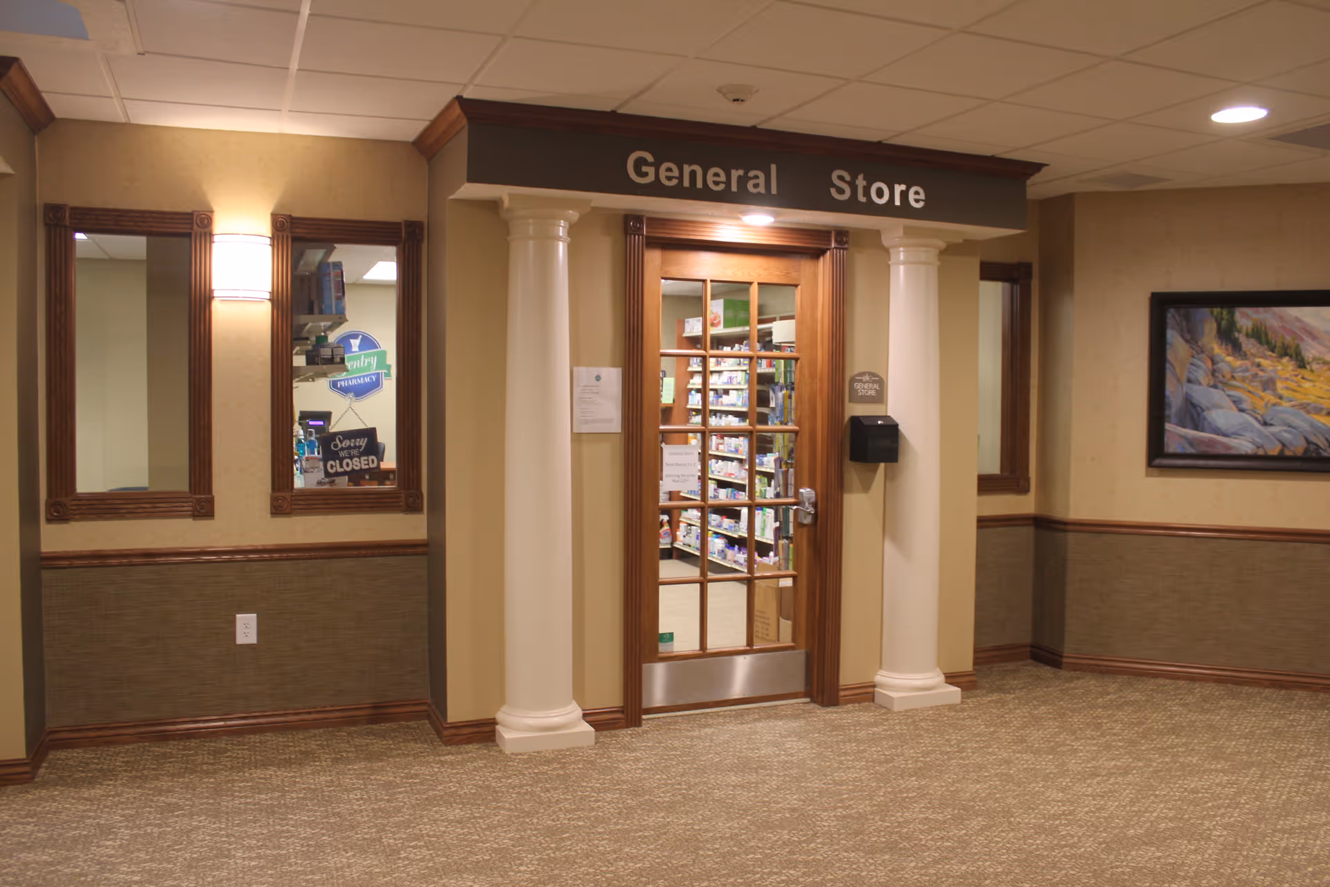 Interior view of a hallway in a senior living facility showing the entrance to a General Store with glass-paneled wooden doors flanked by two white columns. The store shelves inside are stocked with various items. The hallway has beige walls with wooden trim, a carpeted floor, two mirrors on the left wall, a wall-mounted light fixture, and a framed landscape painting on the right wall.