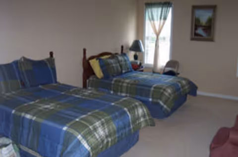 A bedroom with two beds covered in blue and green plaid bedding. There is a window with sheer curtains between the beds, a small table with a lamp, a chair, and a framed picture on the wall.