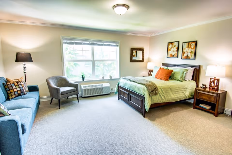 A bright and spacious bedroom in Bridgeport Place Assisted Living featuring a large window with natural light, a wooden bed with green bedding and orange pillows, two bedside tables with lamps, a blue sofa with patterned cushions, a gray armchair, and framed floral artwork on the wall.