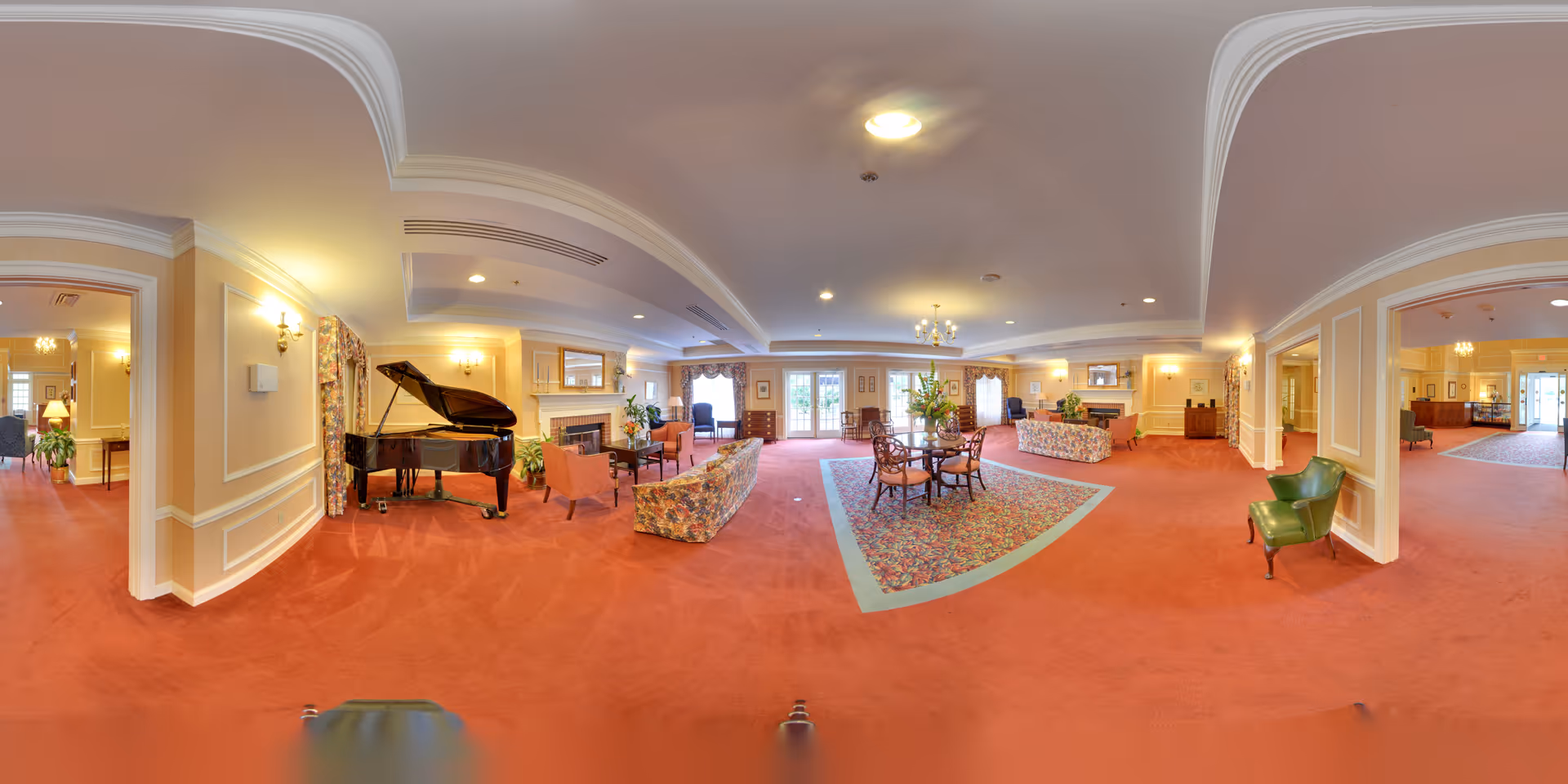 A spacious and elegant senior living facility common area with red carpet flooring, floral patterned sofas, a grand piano, multiple seating arrangements including chairs and small tables, chandeliers, and large windows allowing natural light. The room has cream-colored walls with decorative molding and several plants adding greenery.