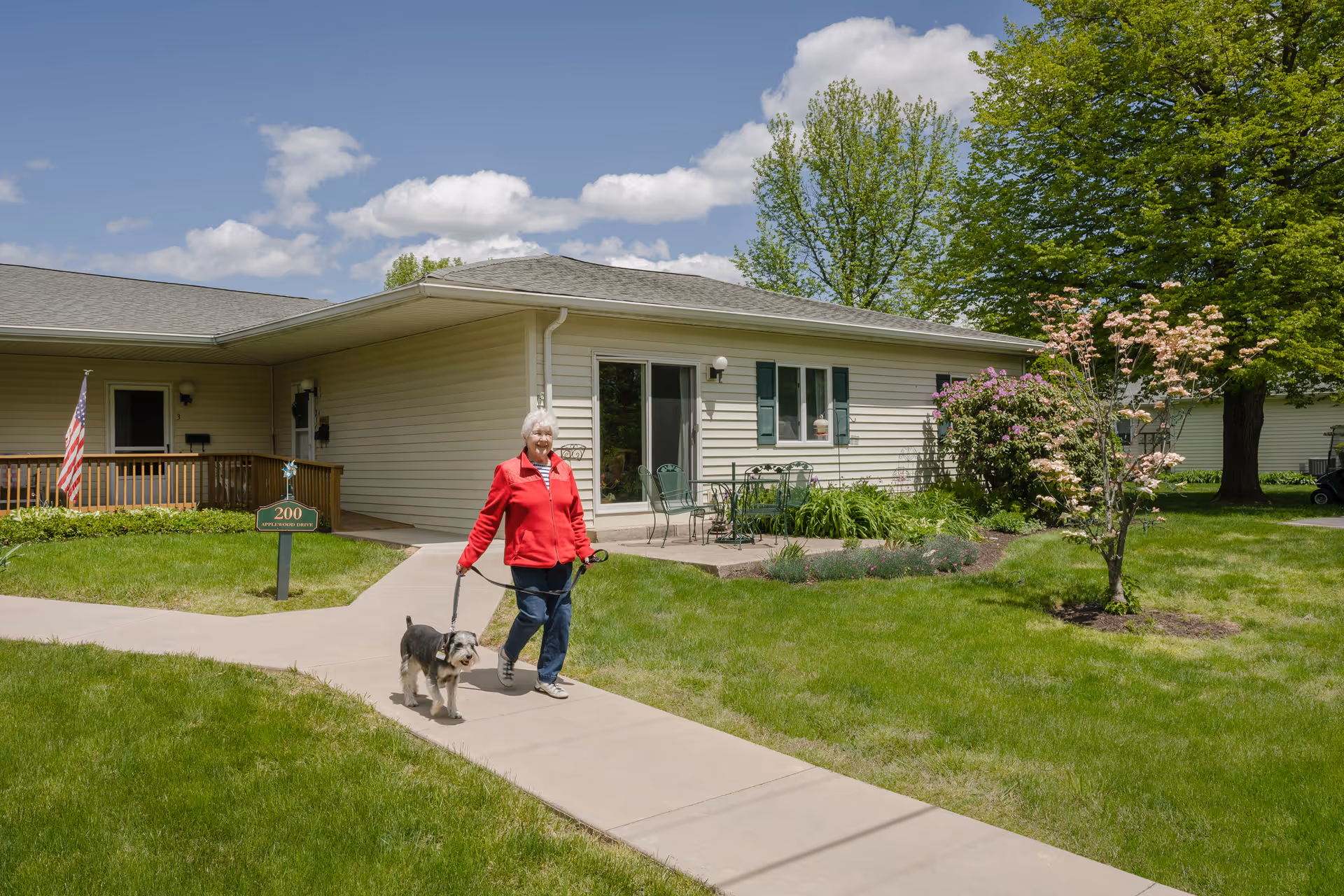 An elderly woman wearing a red jacket walking a small dog on a leash along a paved sidewalk in front of a single-story residential building with beige siding, green shutters, and a small patio with chairs. The scene is set on a sunny day with green grass, trees, and a partly cloudy blue sky.