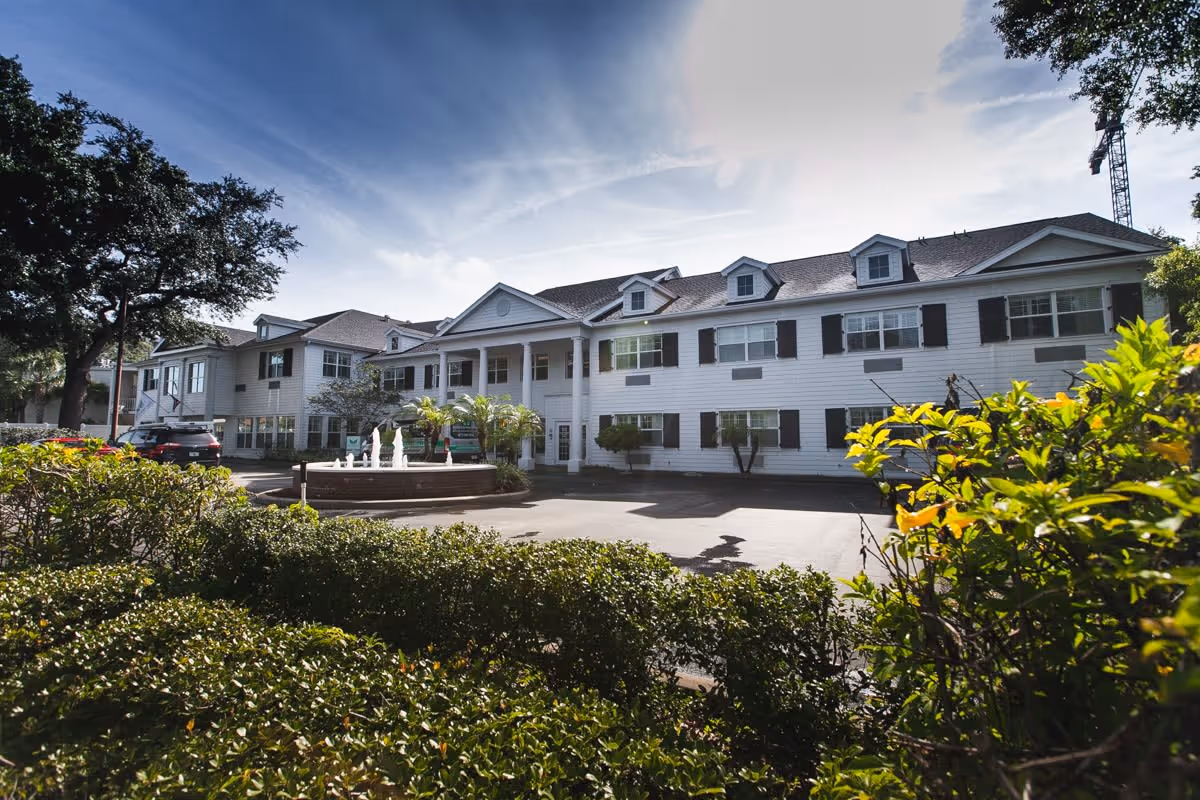 Exterior view of a two-story white senior living facility building with black shutters, a central entrance with columns, a fountain in front, surrounded by greenery and trees under a partly cloudy sky.