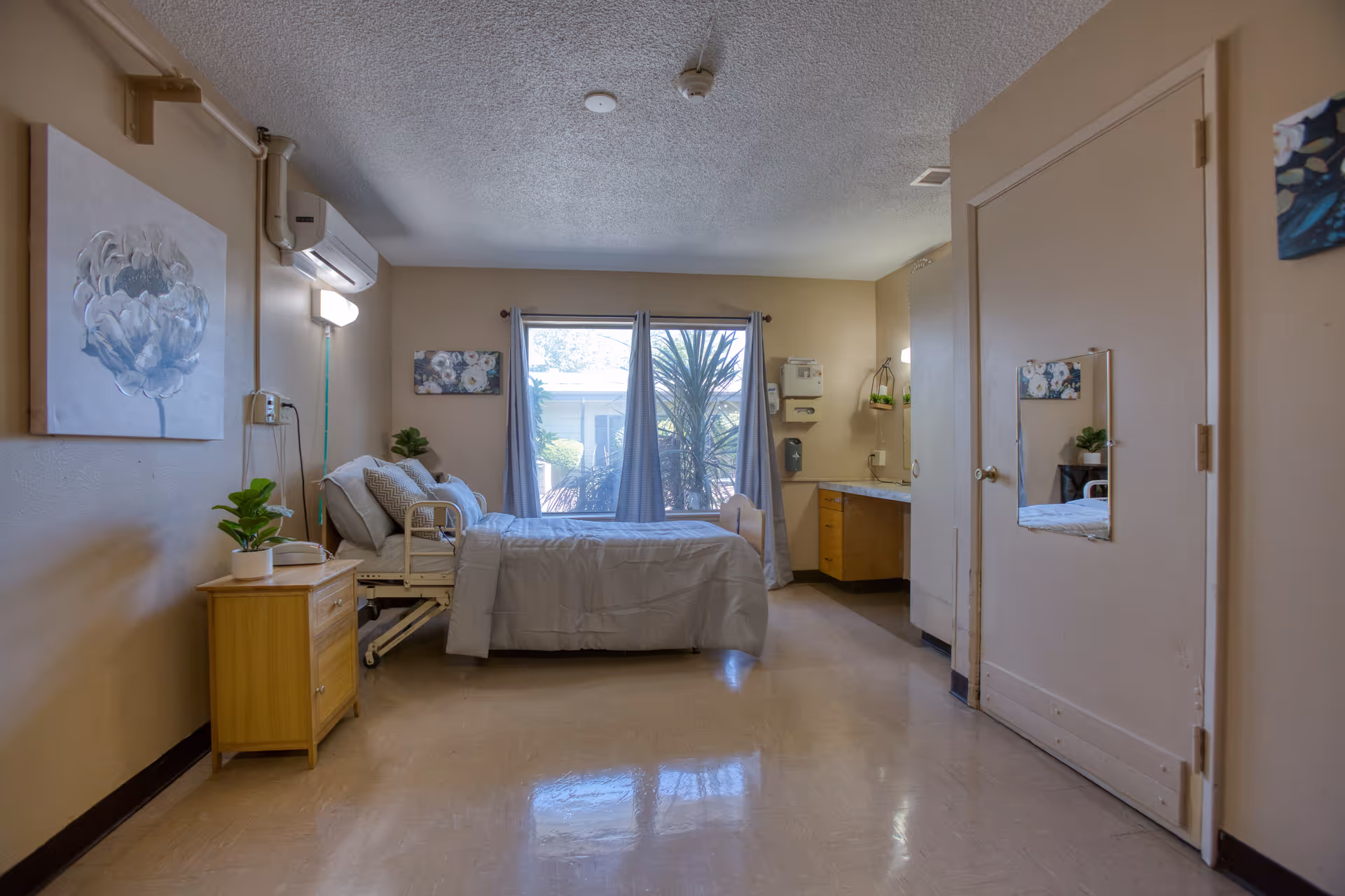 A simple, clean bedroom in a senior living facility with a single bed made up with gray bedding, a wooden nightstand with a potted plant and a telephone, a large window with curtains letting in natural light, a wall-mounted air conditioning unit, and a small desk area with cabinets. The walls are beige and decorated with floral artwork.