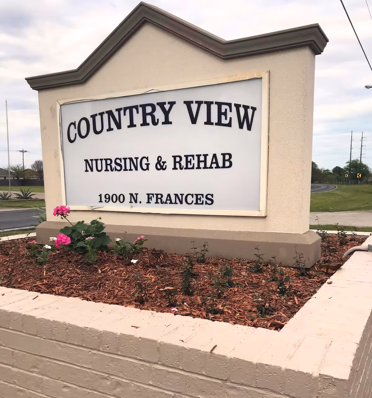 Outdoor sign for Country View Nursing & Rehab located at 1900 N. Frances, surrounded by a small garden bed with mulch and pink flowers, with a road and grassy area in the background under a cloudy sky.