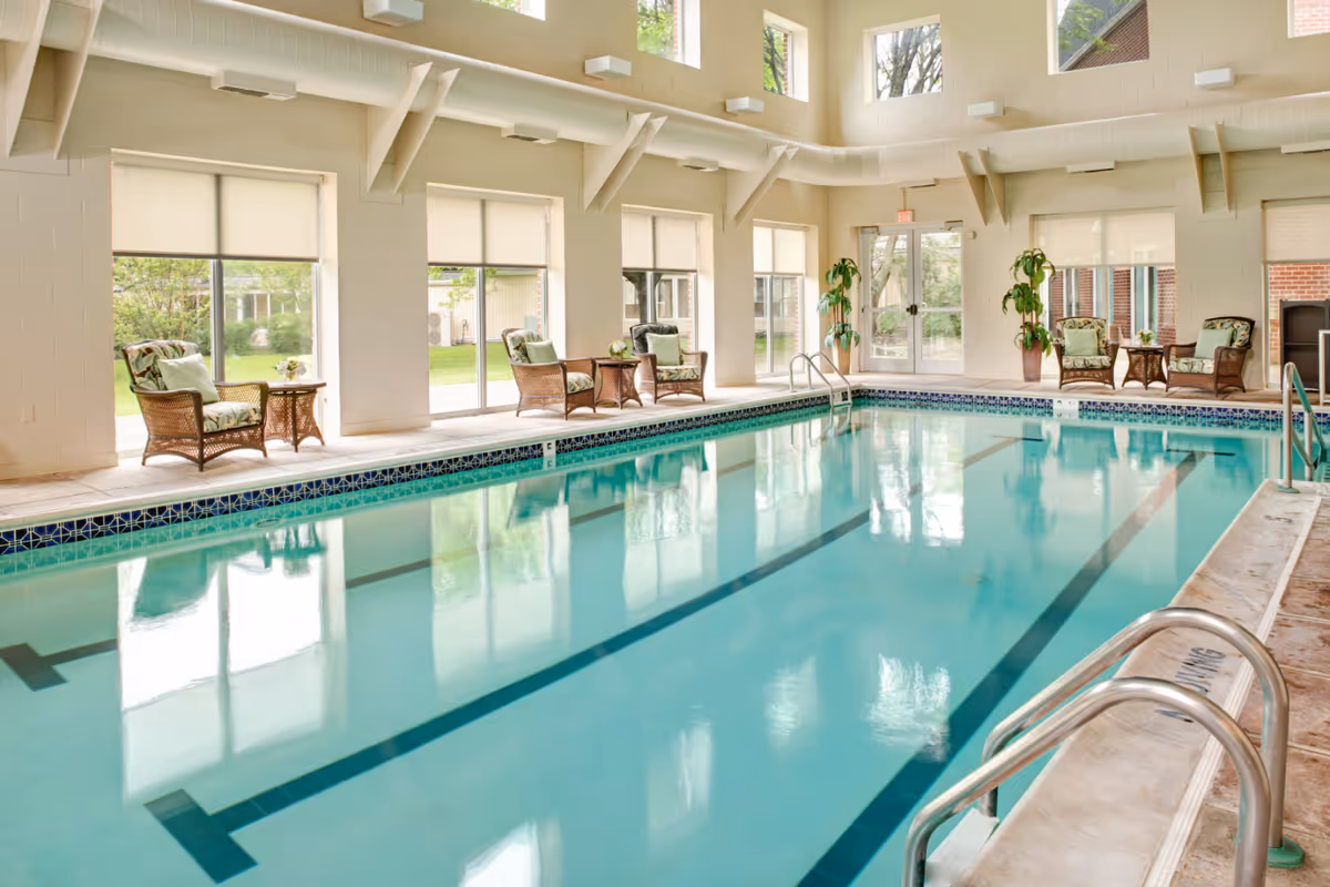 Indoor lap pool in a bright room with wicker lounge chairs, large windows, and potted plants.