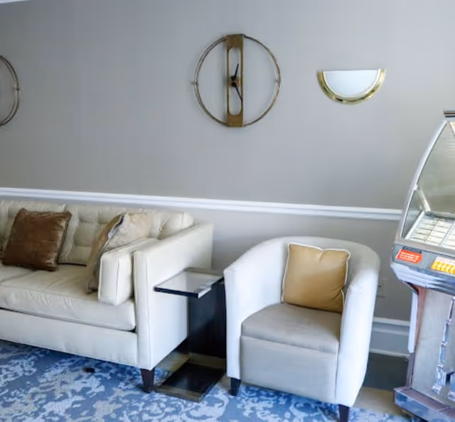 A cozy seating area with a beige sofa adorned with brown and beige pillows, a white armchair with a tan pillow, a small side table between them, a decorative wall clock, a wall sconce, and part of a vintage jukebox on the right side.