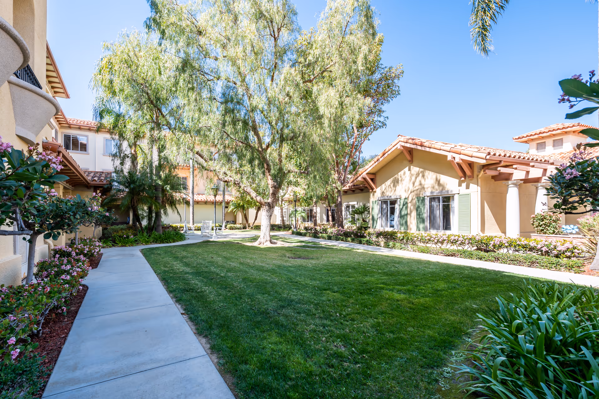 Sunlit courtyard with a central lawn, paved walkways, trees and surrounding single-story residential buildings.