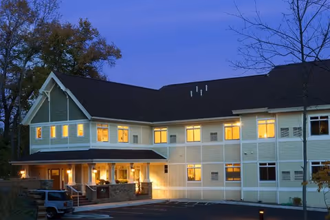 Exterior view of a two-story assisted living facility building at dusk with lights glowing from the windows. The building has a light-colored facade with a dark roof and a covered entrance. There are a few trees around and a parking area in front with a vehicle parked.