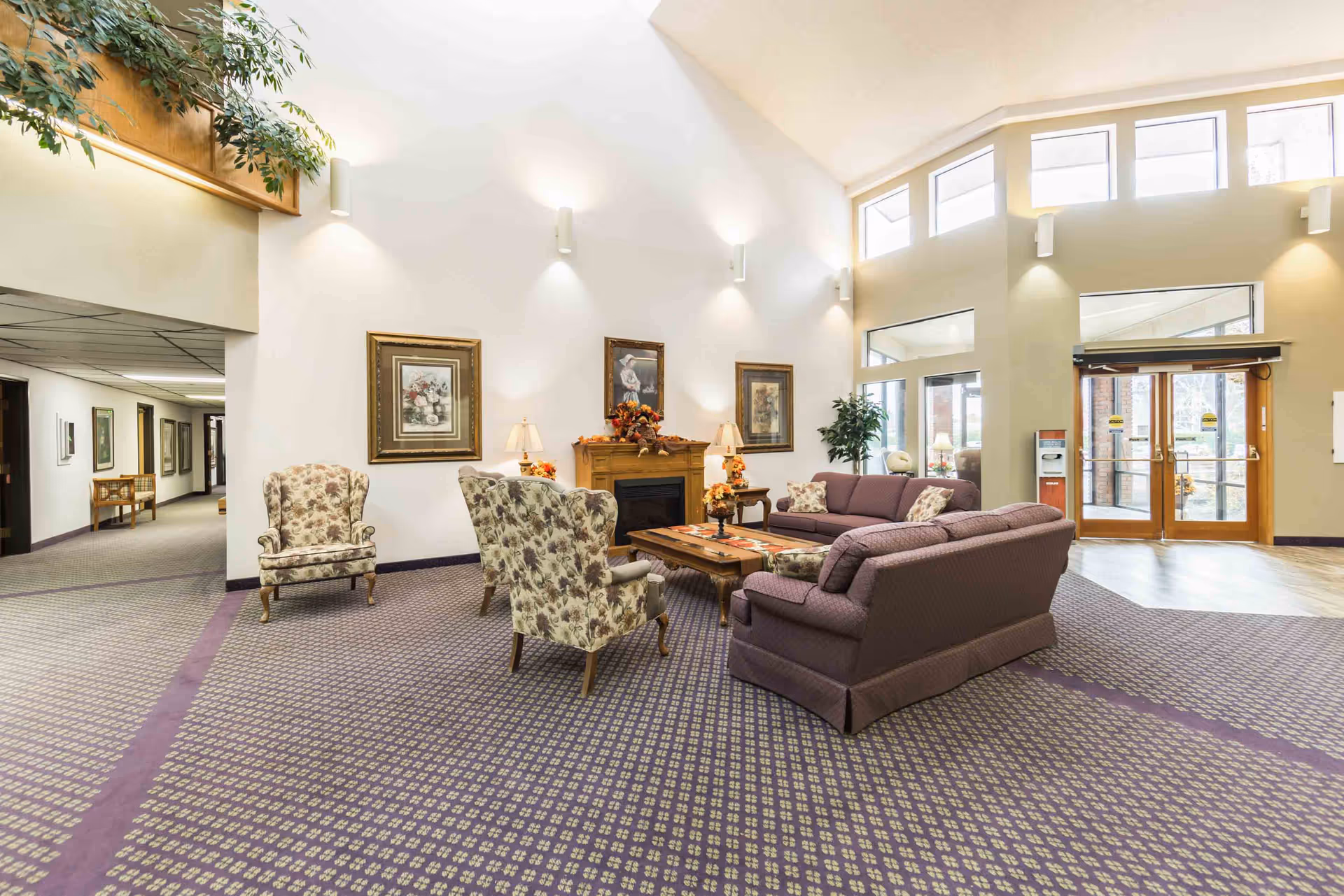 A spacious and well-lit common area with high ceilings featuring a seating arrangement that includes two floral armchairs, two purple sofas, and a wooden coffee table with a decorative runner. The room has a patterned carpet, framed artwork on the walls, a fireplace with autumn-themed decorations, and large windows near the entrance doors allowing natural light to fill the space.