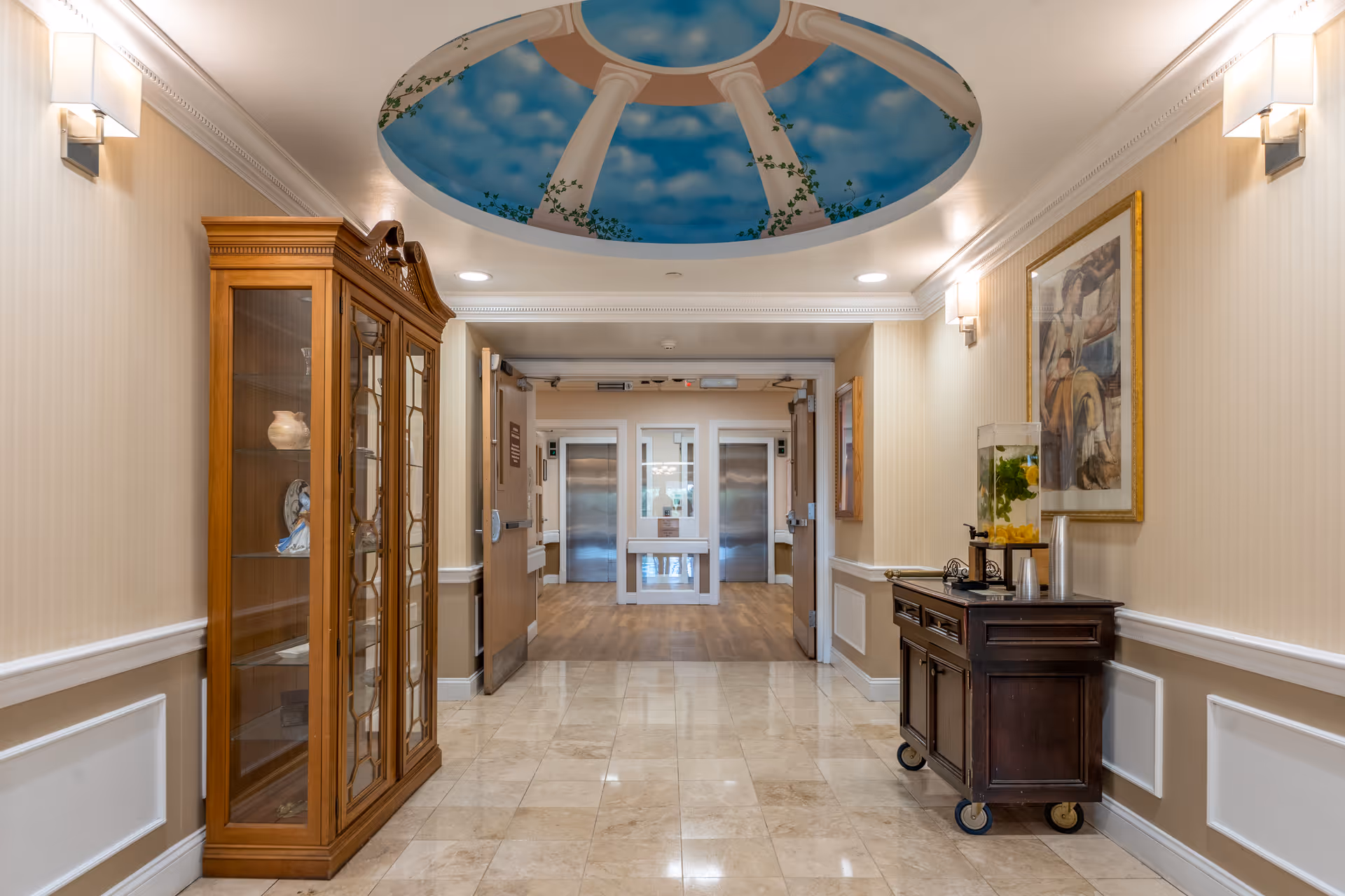 A well-lit hallway in a senior living facility with beige walls, a polished tile floor, and a decorative ceiling mural depicting a blue sky with clouds and white columns. On the left side, there is a wooden glass-front cabinet displaying decorative items. On the right side, a dark wooden cart holds a beverage dispenser with lemon water and cups. At the end of the hallway, double doors lead to an area with two elevators.