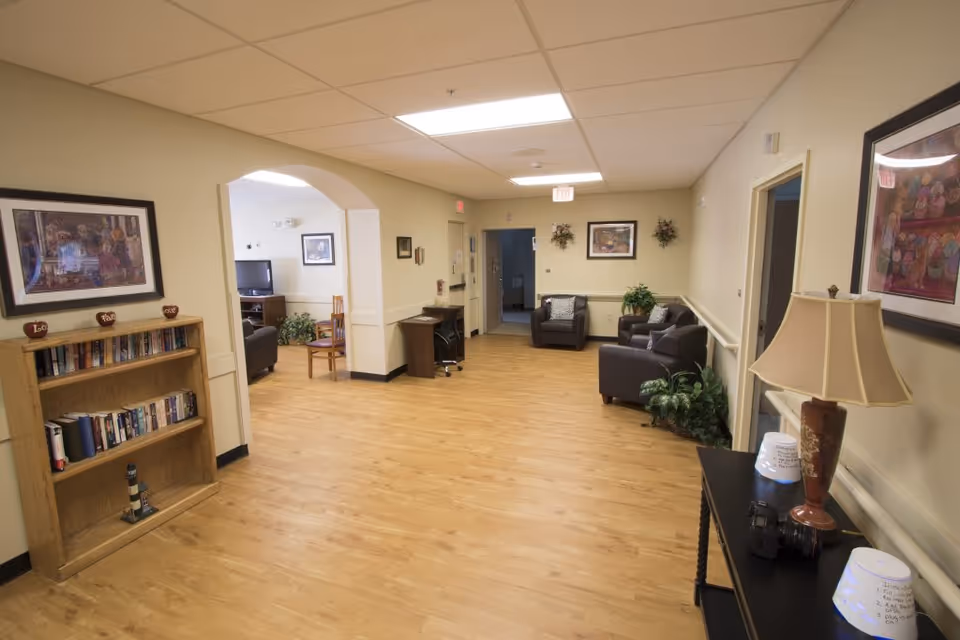 A spacious, well-lit common area in a senior living facility with wooden flooring, beige walls, and ceiling lights. The room features a small bookshelf with books and decorative items, a black table with a lamp and camera, and several armchairs arranged for seating. There are framed pictures and plants decorating the walls and corners. An archway leads to another room with a TV and additional seating.