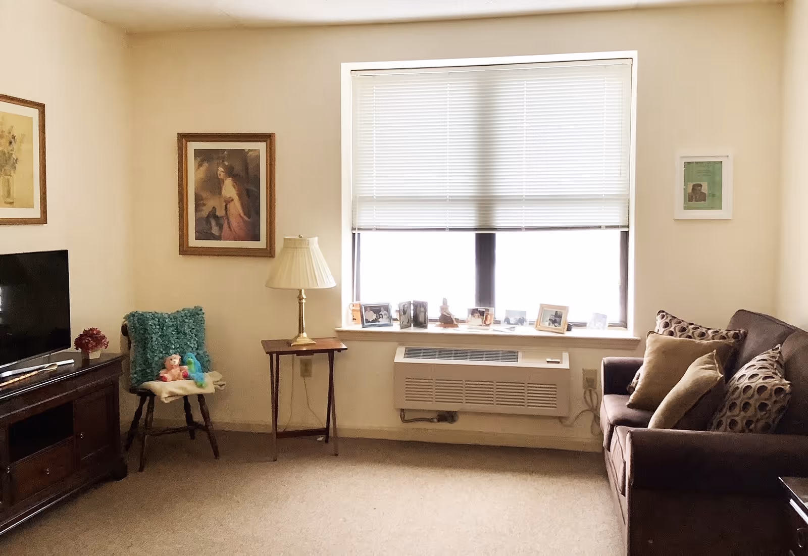 A cozy living room with a brown sofa adorned with patterned and solid pillows on the right. A window with closed blinds is centered on the back wall, with several framed photos and small decorative items on the windowsill. Below the window is a wall-mounted air conditioning unit. To the left, there is a wooden side table with a lamp and a chair with a teal cushion and stuffed toys. A wooden TV stand with a flat-screen television and a small flower arrangement is on the far left. The walls are decorated with framed artwork.