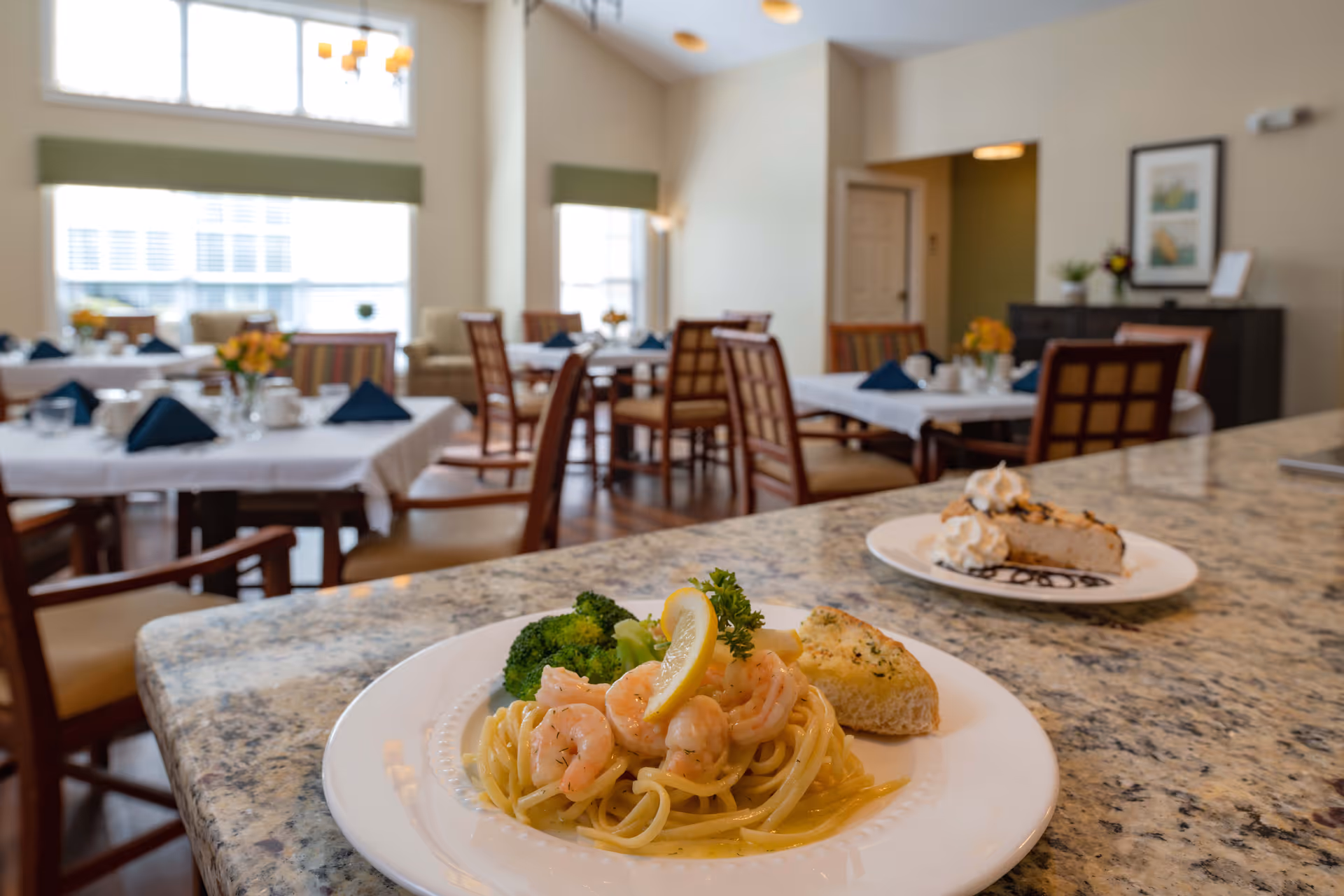 Shrimp pasta and dessert plated on a granite counter with tables set for a bright communal dining room.