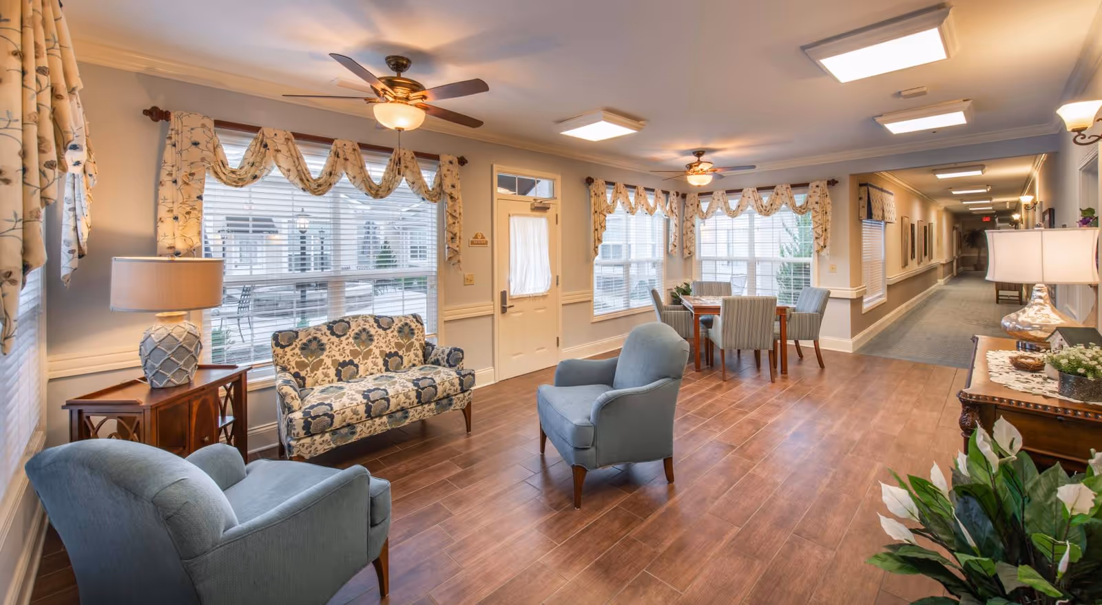 A bright and cozy common area in a senior living facility featuring two blue armchairs, a floral patterned loveseat, a wooden side table with a lamp, and a dining table with four chairs near large windows with floral valance curtains. The room has wood flooring, ceiling fans with lights, and a hallway extending to the right.