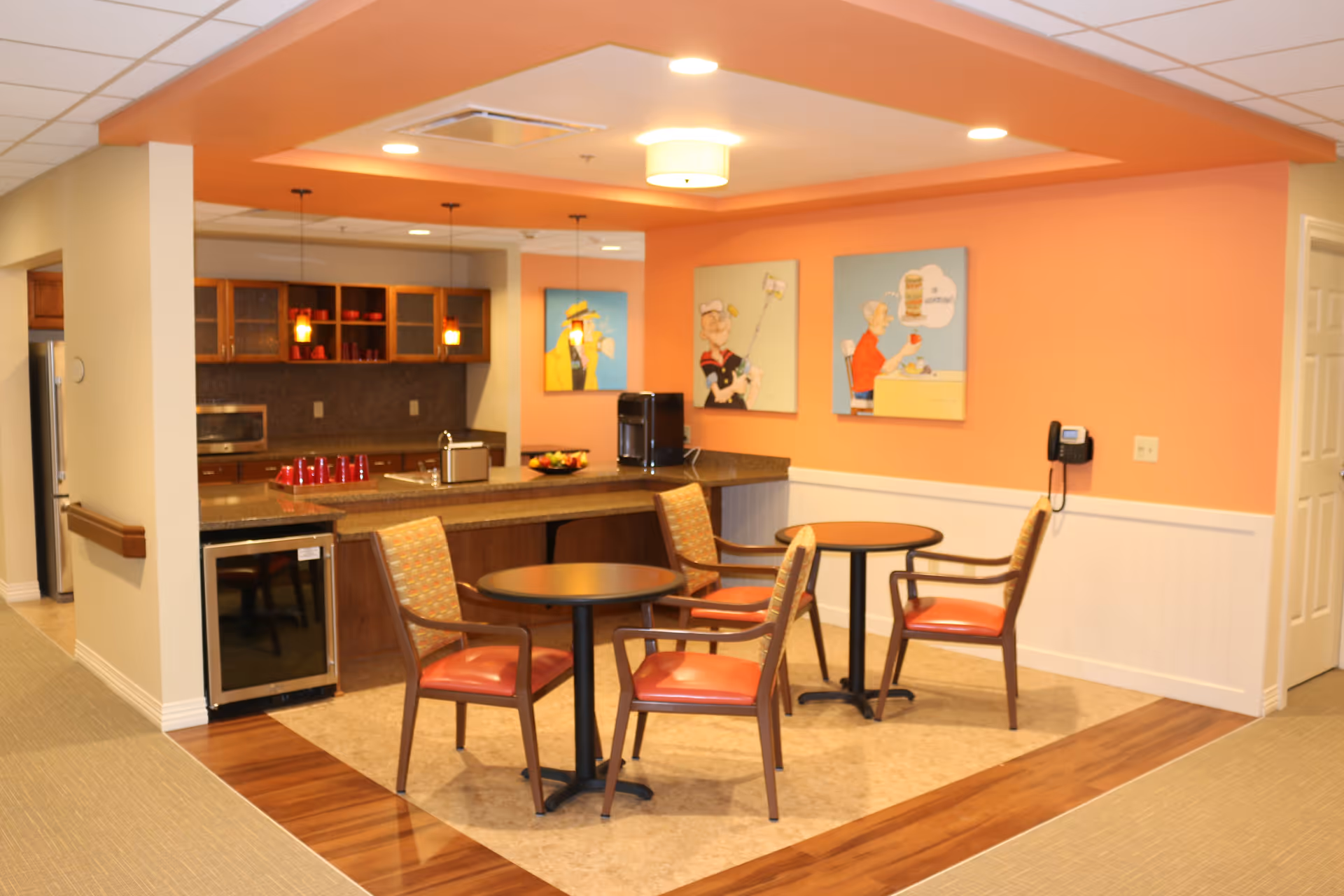 A cozy common area in a senior living facility featuring a small kitchen with wooden cabinets, a countertop with a coffee machine, and two round tables each surrounded by two chairs with orange cushions. The walls are painted peach and decorated with three colorful paintings depicting elderly people. The floor has a combination of carpet and wood-like flooring.