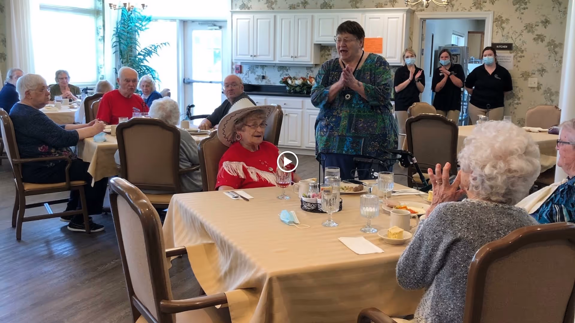 A group of elderly residents seated around tables in a dining room at a senior living facility. A woman stands and claps while others sit and watch. Three staff members wearing masks stand near a doorway labeled 'Kitchen' in the background. The room is well-lit with large windows and decorated with light-colored walls and furniture.