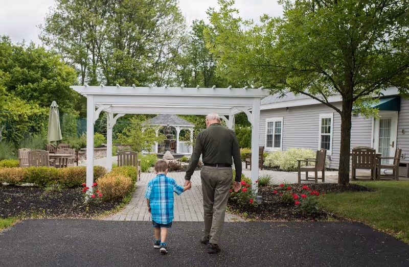 An elderly man and a young boy holding hands and walking on a paved path in a garden area with greenery, flowers, outdoor seating, and a white pergola structure. A gray building with windows and doors is visible on the right side.