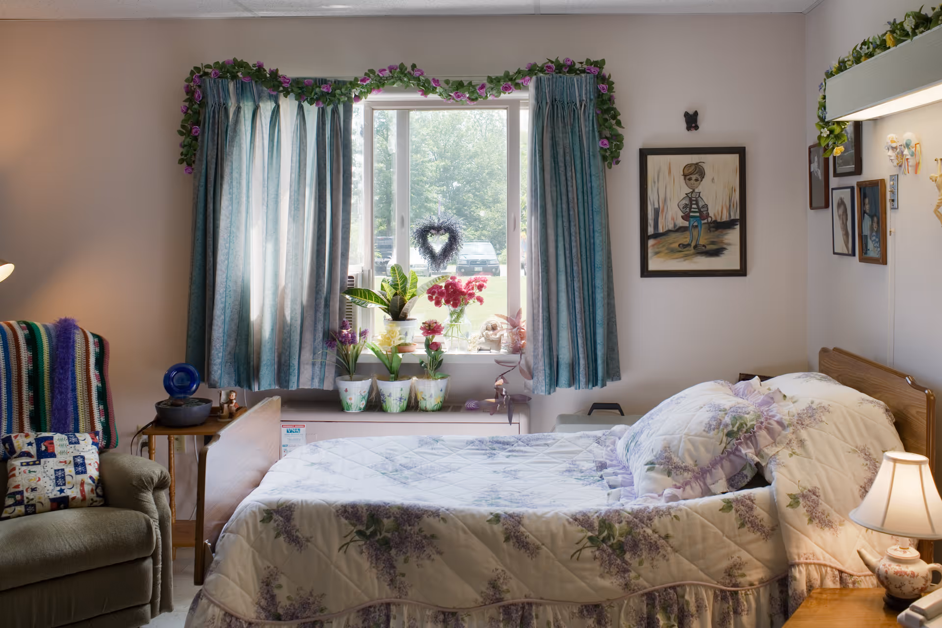 A cozy bedroom with a single bed covered in a floral quilt and matching pillow. Next to the bed is a wooden nightstand with a small lamp. A window with blue curtains and a garland of purple flowers above it lets in natural light. Several potted plants and flowers are placed on the windowsill. To the left, there is a green armchair with a colorful striped blanket and a patterned pillow. The walls are decorated with framed pictures and a small butterfly decoration.