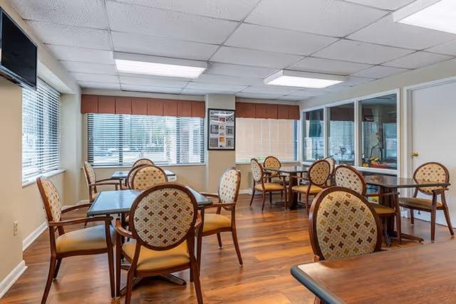 A bright dining room with several wooden tables and cushioned chairs arranged neatly. Large windows with blinds allow natural light to fill the room. The floor is wood, and the ceiling has recessed lighting panels. A television is mounted on the wall near the windows.