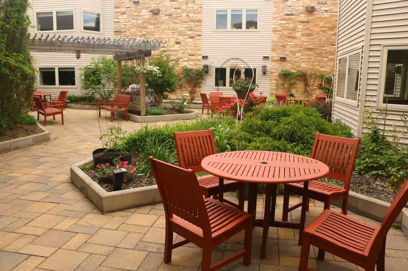 Outdoor courtyard area with red wooden chairs and round tables on a paved surface, surrounded by greenery and plants, with beige building walls in the background.