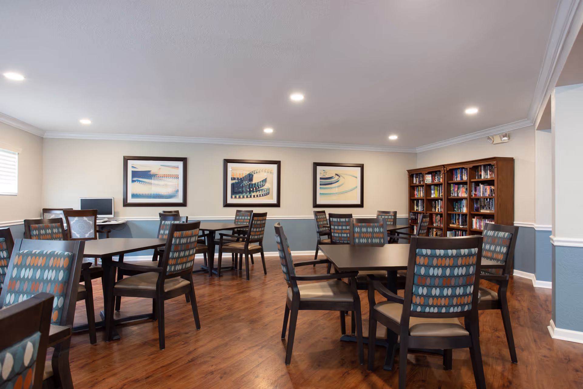 A well-lit room with multiple square tables and patterned chairs arranged neatly on a wooden floor. The walls are painted beige with a blue wainscot and decorated with three framed abstract art pieces. There is a bookshelf filled with books in the corner and a computer on a small desk near a window with blinds.