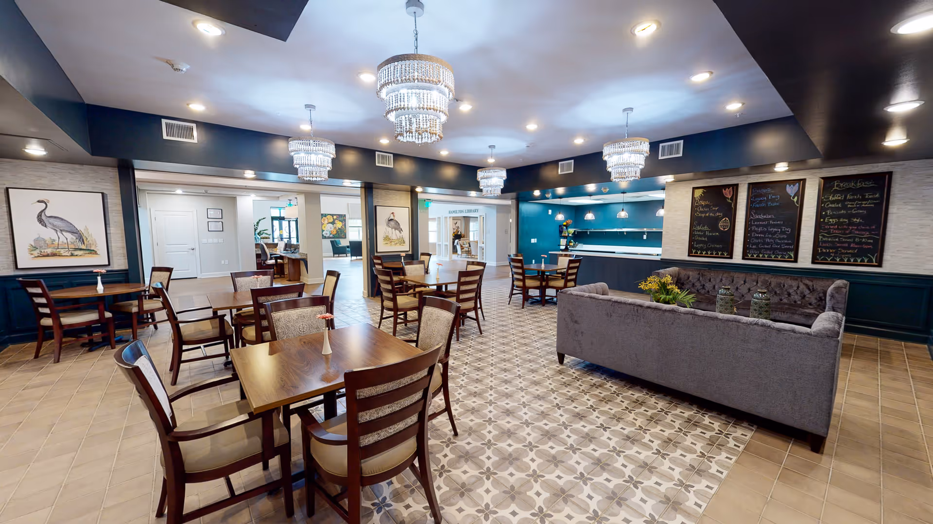 A spacious dining room featuring wooden tables and chairs, elegant chandeliers, and decorative artwork on the walls. The room has a cozy atmosphere with a gray sofa and a chalkboard menu in the background.